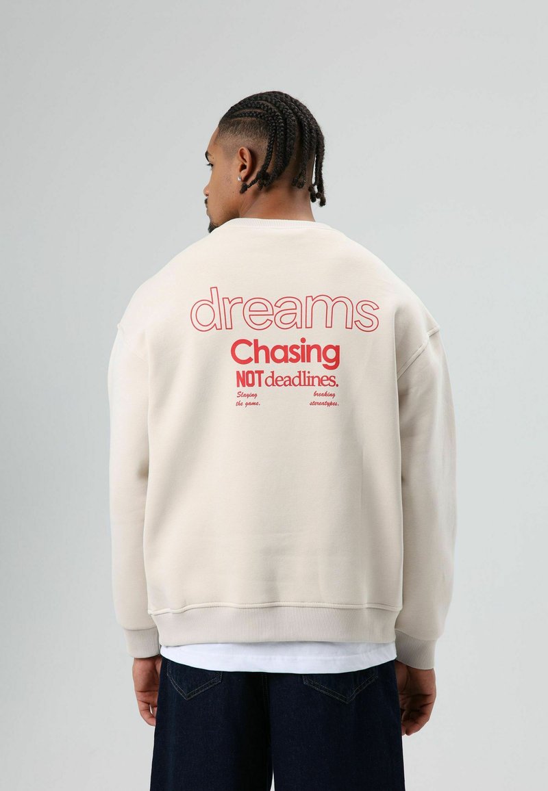 Beige sweatshirt met rode tekst op de achterkant: "dreams Chasing NOT deadlines." Oversized pasvorm met geribde boorden en zoom.