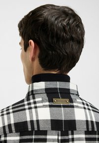 Zwart-witte geruite shirt met een ronde kraag, voorzien van een geweven label aan de achterkant. De stof lijkt textuur te hebben en is met knopen vastgemaakt.