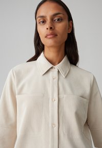 Camicia di velluto a coste beige chiaro con colletto a punta, chiusura a bottoni e due tasche sul petto. Il tessuto presenta un motivo a righe in rilievo.