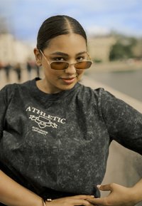 Schwarzer Sweatshirt mit marmoriertem Muster und weißem Text "ATHLETIC ELITE LEAGUE UNIVERSITY" sowie kleinem Logo. Das Modell trägt goldgetönte Sonnenbrillen.