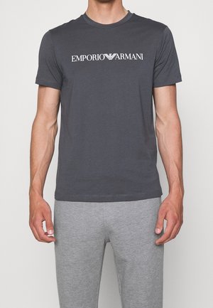 Homme portant un t-shirt Emporio Armani gris foncé et un pantalon gris clair, debout devant un fond clair uni.