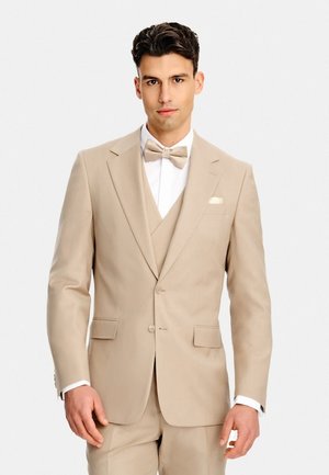 Młody mężczyzna w beżowym trzyczęściowym garniturze z pasującą muszką, stojący na tle jednolitego białego tła.