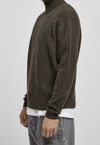 Maglione lavorato a maglia marrone scuro con colletto alto, polsini e orlo a costine. Texture morbida, vestibilità regolare, indossato sopra una maglietta bianca.