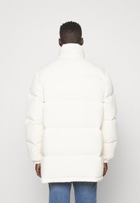 Bílá péřová bunda s vysokým límcem, zatepleným designem a viditelným prošíváním. Měkká textura a široký silueta pro pohodlí.