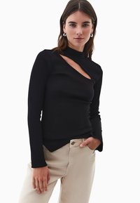 Czarna bluzka z długim rękawem, o wysokim dekolcie i wycięciu. Żebra tekstura, dopasowany krój, zestawiona z jasnobeżowymi spodniami.