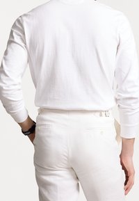 Långärmad vit bomullsskjorta i kombination med vita byxor, med bakfickor och detaljer för bälteshällor. Slät textur, minimalistisk design.