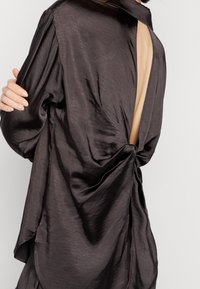 Haut gris foncé en tissu soyeux avec un design drapé. Présente un twist dans le dos et un dos ouvert pour un effet superposé. Manches longues et coupe décontractée.