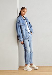 Lichtblauwe denimjas met een oversized fit, gecombineerd met lichtblauwe denimjeans met zijstrepen. Witte sneakers met beige accenten.