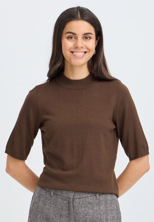 Femme souriante aux longs cheveux foncés, portant un haut en maille marron à manches courtes et un pantalon gris, debout devant un fond clair.