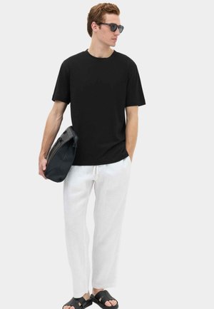 Junger Mann trägt schwarzes T-Shirt, weiße Hose, schwarze Sandalen und Sonnenbrille, hält eine schwarze Tasche und schaut zur Seite.