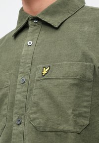 Camisa verde texturizada con diseño de botones, dos bolsillos frontales y un logo amarillo bordado en el pecho.