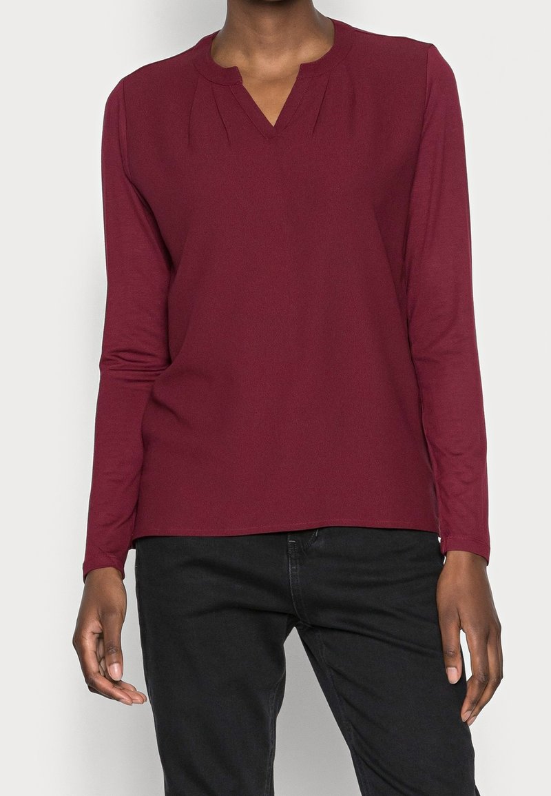 Personne portant un haut bordeaux à manches longues avec des détails plissés près du col et un pantalon noir, debout devant un fond uni.