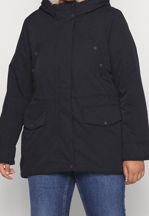 Personne portant une veste d'hiver noire à capuche avec poches avant à rabat sur un jean bleu sur un fond uni.