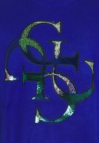 Niebieski dzianinowy top z teksturowanym wzorem z zielonych i srebrnych cekinów, przedstawiającym splecione litery w odważnym, błyszczącym wzorze.