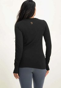 Svart långärmad skjorta med åtsittande design, rund hals, och broderad logotyp vid bakre nacken, kombinerad med grå leggings. Slät yta.