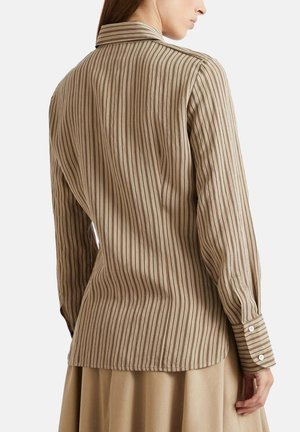 Vrouw die een beige shirt met lange mouwen en donkere verticale strepen draagt, met haar rug naar de camera, gecombineerd met een beige rok.