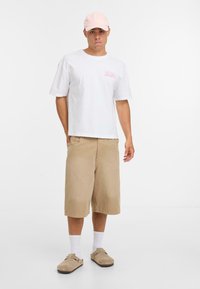 Camiseta de algodón blanca con gráfico rosa, pantalones cortos caqui, zuecos beige y gorra rosa clara; diseño limpio, ajuste casual, mangas cortas.