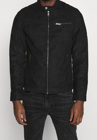 Chaqueta de ante sintético negro con cierre de cremallera, bolsillos laterales y un bolsillo en el pecho. Presenta un cuello alzado y detalles de costura en los hombros.