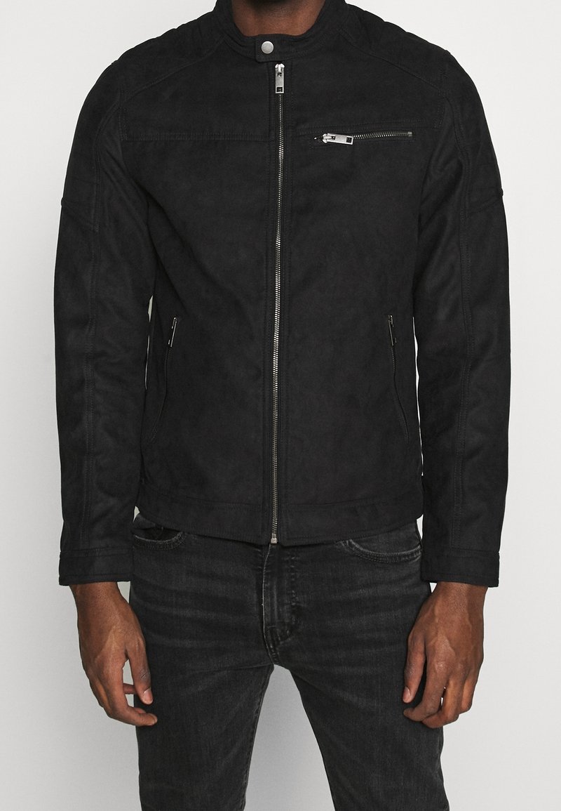 Chaqueta de ante sintético negro con cierre de cremallera, bolsillos laterales y un bolsillo en el pecho. Presenta un cuello alzado y detalles de costura en los hombros.