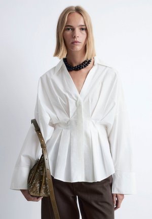 Hvid langærmet skjorte med V-udskæring, med plisseringer og en afslappet silhuet. Kombineret med mørkebrune bukser og en vævet brun håndtaske.
