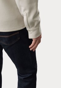 Sudadera beige con detalles en los puños, combinada con unos pantalones vaqueros oscuros. El puño presenta un acento de botón y la tela tiene una textura suave.