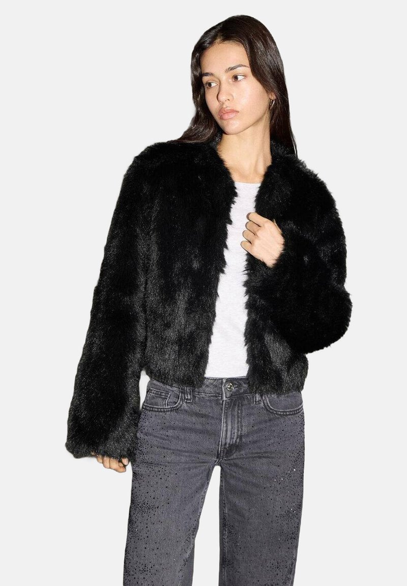 Schwarze Kunstfelljacke mit einem kurzen Design und weiten Ärmeln, getragen über einem weißen Oberteil und kombiniert mit dunklen Jeans mit dezenten Verzierungen.