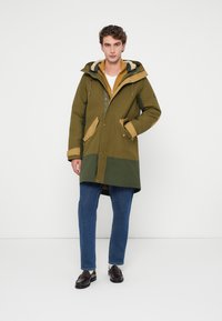 Parka vert olive avec des touches de jaune et de vert foncé, dotée d'une capuche, d'une fermeture éclair et de poches avant. Associée à un jean bleu et des chaussures foncées.