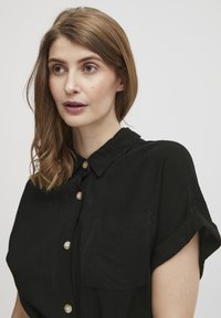 Cămașă neagră cu guler, mâneci scurte răsucite și un buzunar frontal, având nasturi de culoare deschisă și un material texturat.
