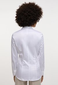 Camicia bianca a maniche lunghe con colletto classico, tessuto morbido, dettagli di cucitura sulla schiena e polsini con bottoni. Design pulito e sartoriale.