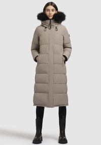 Kvinde iført en lang beige dunjakke med sort pelskrave, sorte leggings og sorte kraftige støvler, stående med hænderne i lommerne.