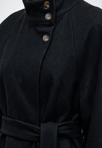 Manteau en laine noire avec col haut, trois boutons visibles et une ceinture nouée à la taille, montré de la poitrine jusqu'à la hanche.