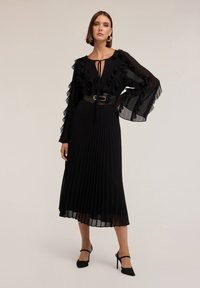 Schwarzes Midi-Kleid mit Rüschensäumen, plissiertem Rock und einem glänzenden schwarzen Gürtel. Es hat einen tiefen Ausschnitt mit einer Bindeschließe. Schwarze High Heels.