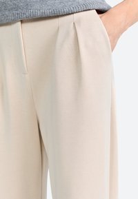 Beige broek van zachte stof, met voorplooien en zijzakken. De gladde textuur draagt bij aan een verzorgde uitstraling.