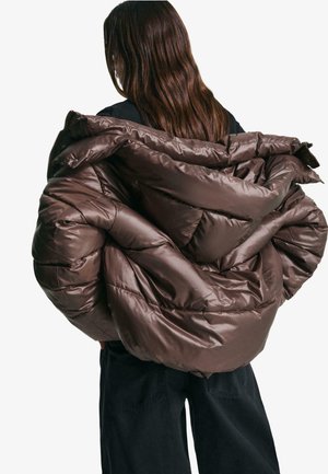 Bruine opvouwbare pufferjack met een glanzende afwerking, oversized ontwerp, gequilt textuur en een capuchon, gedragen over een zwart top.