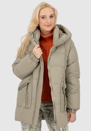 Steppjacke in hellem Olivgrün mit Steppmuster, Reißverschluss und Druckknöpfen, Seitentaschen, kombiniert mit einem grob gestrickten orangefarbenen Pullover.