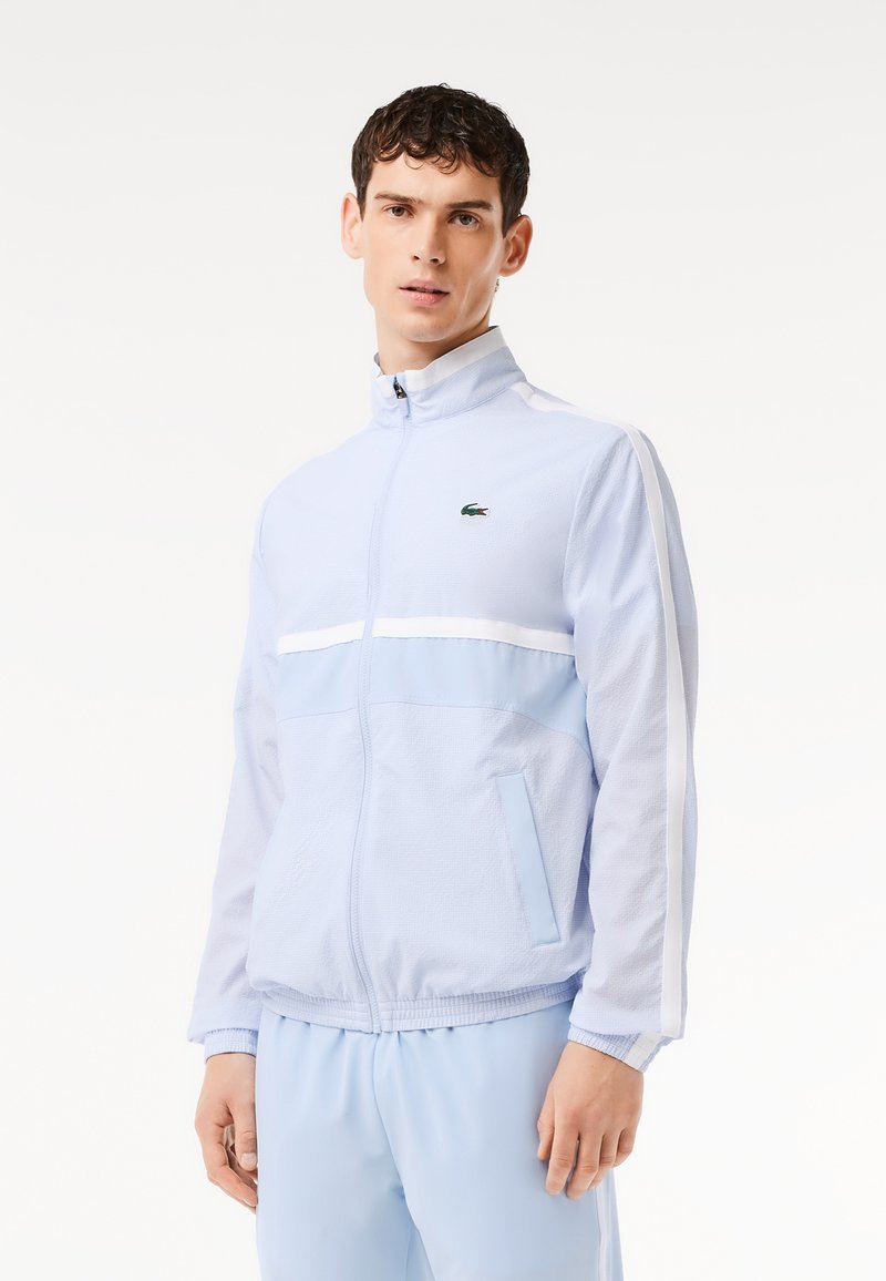 Veste zippée bleu clair en tissu texturé, avec une bande blanche sur la poitrine et un logo vert brodé sur le côté gauche.