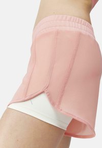 Svetlo roza atletske kratke hlače iz zračnega materiala s koničastim robom, položene čez bele notranje hlače. Vidni šivi okrasijo šive.