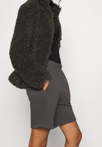 Veste en fausse fourrure noire, texturée et offerte, portée sur un top noir à côtes. Associée à un short doux gris, long jusqu'aux genoux, avec des poches latérales.