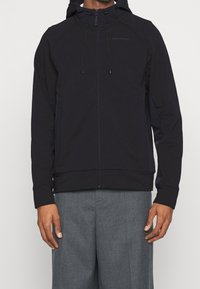Giacca con cappuccio nera e zip completa, cappuccio con cordino e maniche raglan. Presenta un design aderente e una texture di tessuto morbido.