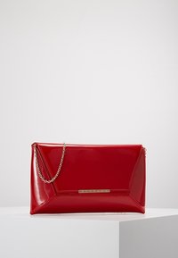 Pochette rectangulaire rouge brillante avec couture géométrique et fine chaîne en or sur une surface blanche.