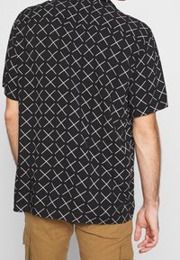 Schwarzes T-Shirt mit kurzen Ärmeln und einem weißen diagonalen Kreuzmuster, hergestellt aus leichtem Stoff mit lockerer Passform und Kragen.