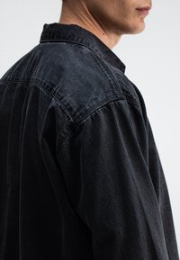 Zwart denim overhemd met een ontspannen pasvorm, voorzien van een puntige kraag, knopen op de sluiting en zichtbare stiksels op de schouders.