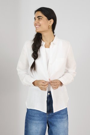 White linen shirt with a collar, button front, long sleeves, and single chest pocket, paired with blue high-waisted jeans.