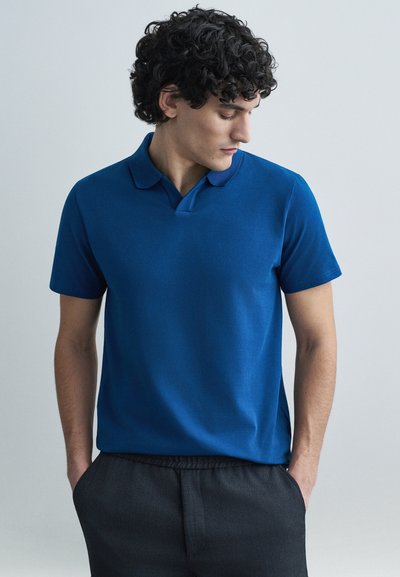 Blå polo-shirt med blød tekstur, korte ærmer og krave. Båret med mørke, elastiske bukser med talje. Ensfarvet design uden mønstre.