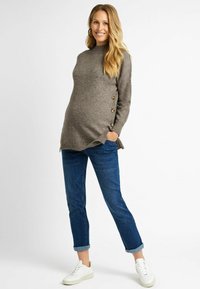 Pull de maternité en tricot marron clair avec un col haut, des boutons sur les côtés et un ourlet incurvé, associé à un jean bleu retroussé et des baskets blanches.