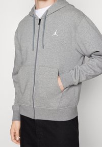 Sudadera gris con cierre de cremallera, hecha de material de sudadera. Cuenta con un bolsillo frontal, capucha con cordón y un logo blanco en el lado izquierdo del pecho.