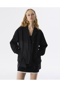 Chaqueta negra con rayas, con un corte relajado, detalles de cordón y dos bolsillos frontales. Lleva puesta sobre una falda negra.