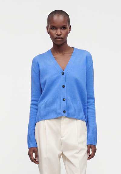 Femme portant un cardigan bleu boutonné avec ourlet et poignets côtelés, associé à un pantalon beige taille haute, se tenant devant un fond uni.
