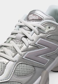 Ασημί και γκρι αθλητικό παπούτσι New Balance 9060 με λευκές κορδόνια και λεπτομέρειες από δίχτυ, εμφανιζόμενο σε κοντινή λήψη από πλευρική γωνία.
