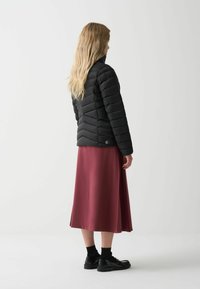 Zwarte pufferjack met een chevron-gequilt patroon, gecombineerd met een lange bordeauxrode rok. Model draagt zwarte schoenen en is van achteren gezien.